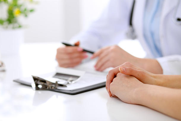 Close-up of patient's hands resting on a desk while a healthcare provider in white coat writes on clipboard in the background during a medical consultation.
