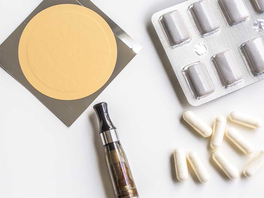 Various smoking cessation products including an e-cigarette device, nicotine patches, pills in blister packs, and white capsules arranged on a white surface.
