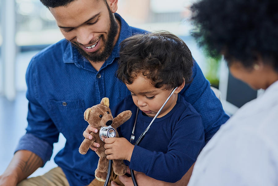 A pediatric patient wears a stethoscope and holds a stuffed bear as his father smiles at him