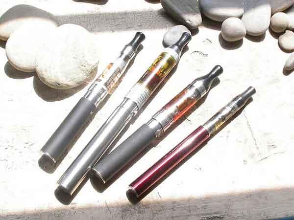 Five e-cigarettes with various colored tanks and cartridges scattered on a light wooden surface.