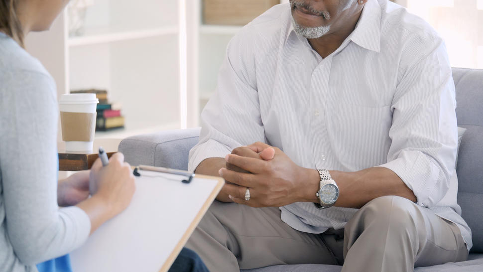 Seated female health care provider with clipboard meets with seated male patient.