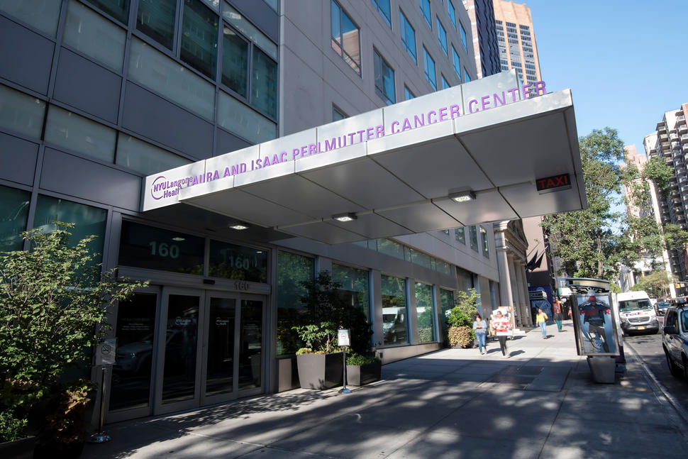 An exterior photo of the Laura and Isaac Perlmutter Cancer Center at NYU Langone Health. Only the lower floors of the building are visible along with the awning of the building that contains a sign with the name of the center. In the background, other tall buildings and trees can be seen along with cars on a street and people walking on a sidewalk. The sky is blue and cloudless.