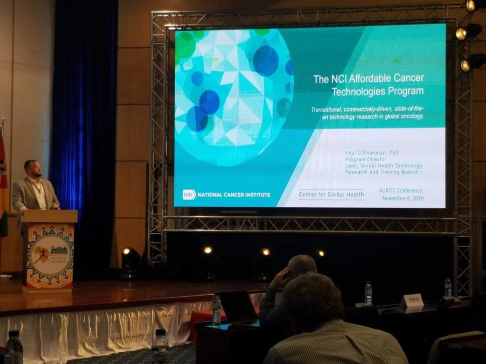 Paul Pearlman stands at a podium on stage and looks at a large screen. He has closely cropped dark hair and beard and wears a suit without a tie. The screen shows the title slide of a presentation: “The NCI Affordable Cancer Technologies Program: Translational, commercially-driven, state-of-the-art technology research in global oncology.” The slide also has the NCI logo, Pauls’ title, “Center for Global Health” and conference name with date of presentation (Nov 6).