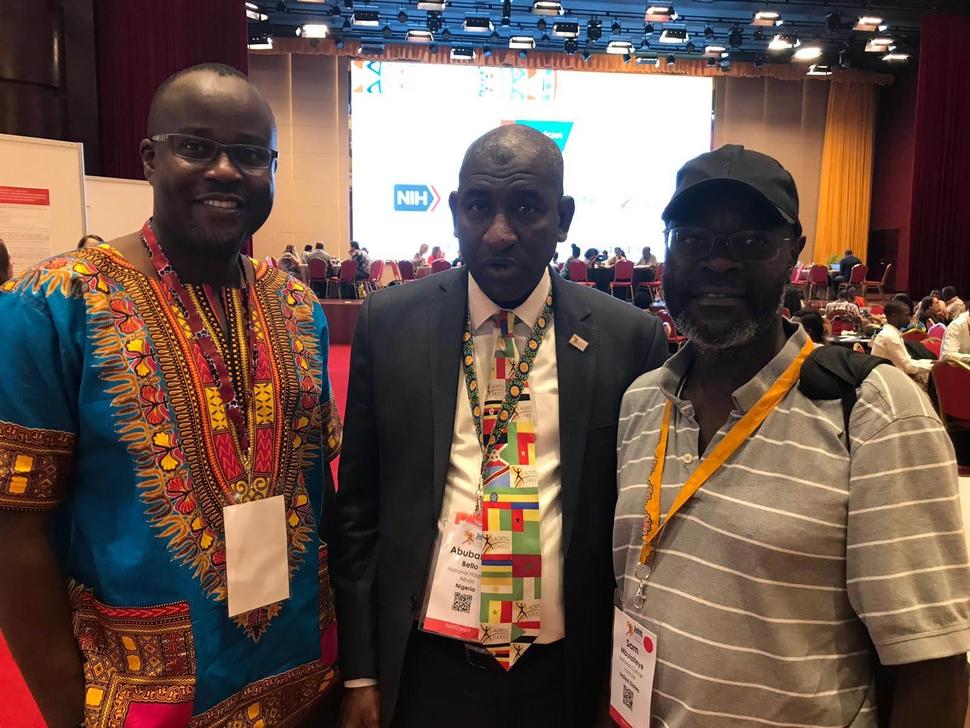 Three Black men stand looking at the camera with a conference hall in the background. The man on the left wears a dashiki shirt, the man in the center wears a suit and tie, and the man on the right wears a hat and short sleeve grey polo shirt. They all wear lanyards around their necks. In the background there’s a large screen with the NIH logo.