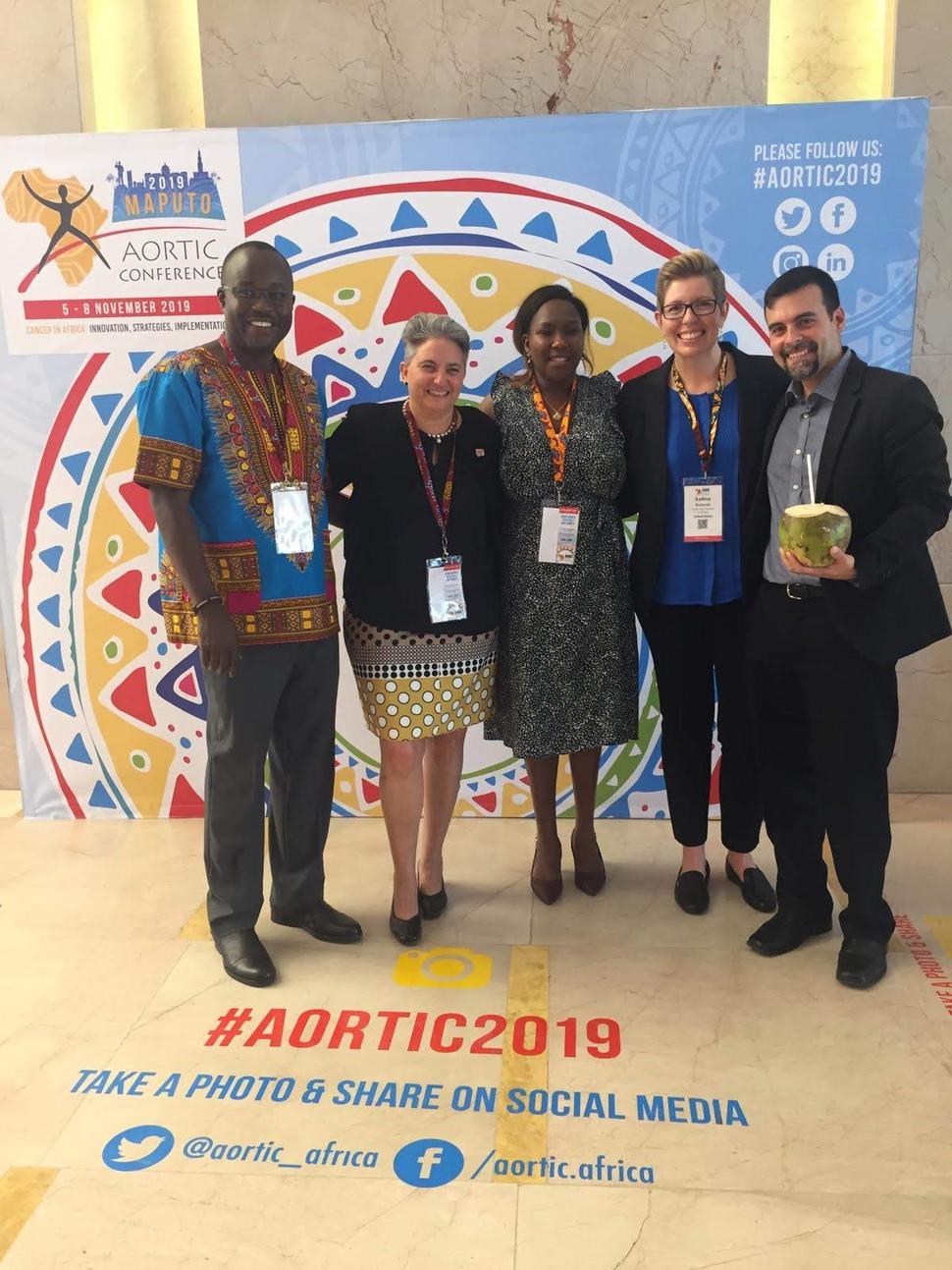 Two men and three women stand looking at the camera, smiling, in front of a backdrop for the Aortic Conference. One man wears a dashiki shirt with slack and dress shoes; everyone else is dressed in business clothing. On the floor in front of the group is the hashtag #AORTIC2019 with Twitter and Facebook contact information.