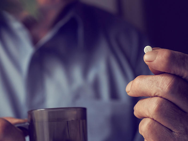 A close up of a hand holding up a small, round, white pill. The hand and pill are in focus. In the background, out of focus, is another hand holding a cup and button-up shirt. Part of the man’s neck can be seen as well.