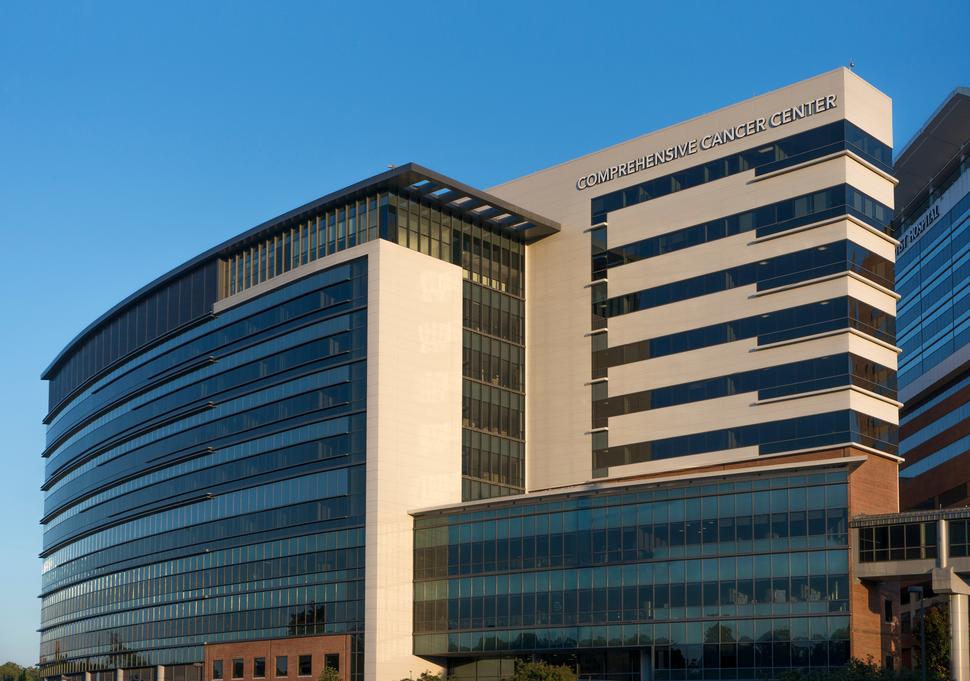 Exterior view of Wake Forest Baptist Comprehensive Cancer Center (WFBCCC). The building is a modern, multi-story building labeled "Comprehensive Cancer Center" and features large glass windows and a sleek, curved design, reflecting the sunlight.