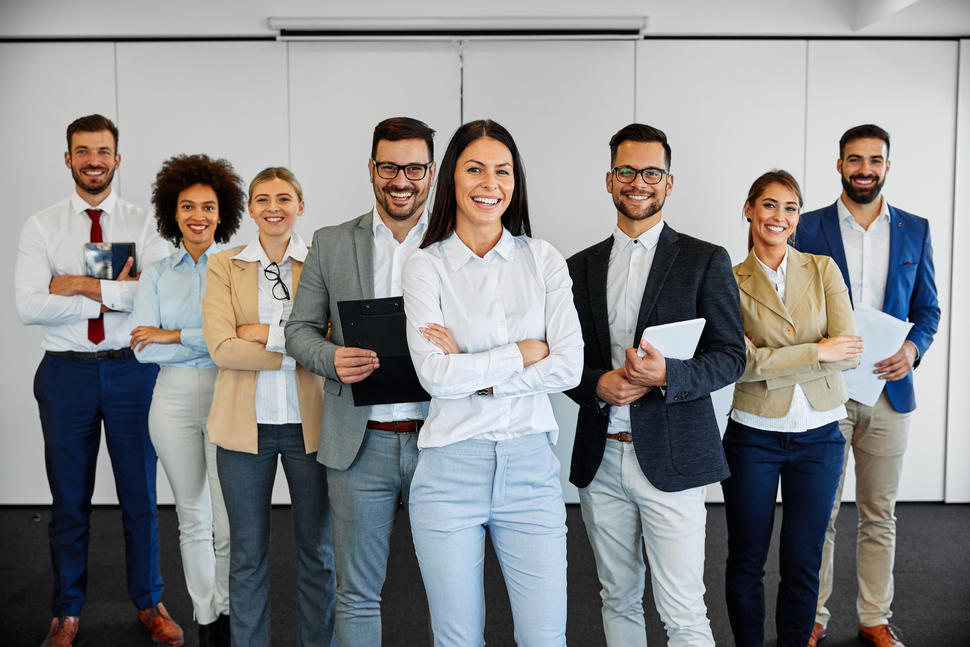 A multi-ethnic group of people, all of whom are dressed in business casual clothing, are lined up in a row and look at the camera, smiling. 