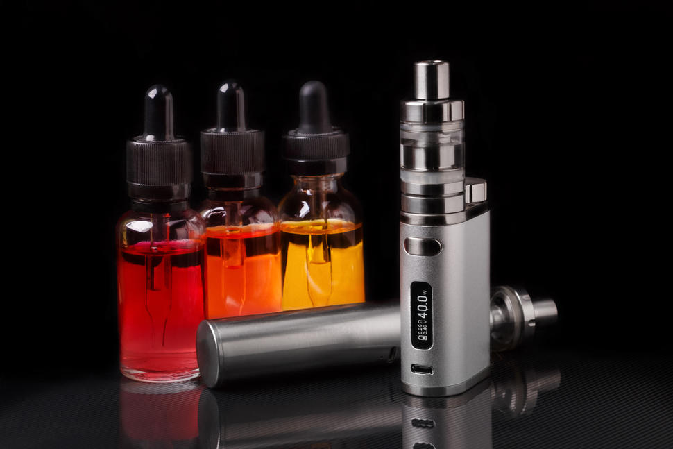 An image of an e-cigarette machine in the foreground with three small bottles that have eyedropper tops in the background. The small bottles contain liquids colored red, orange and yellow; the e-cigarette machine is grey with metal touches.