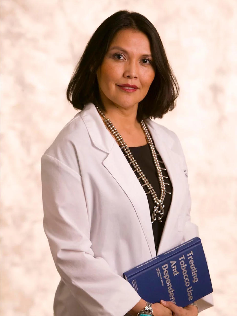 Patricia Henderson stands looking at the camera and wearing a lab coat. She is holding a blue book with the title "Treating Tobacco Use and Dependency".