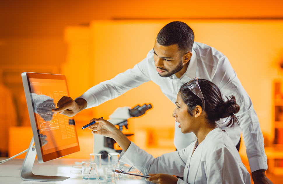 Two scientists at a lab setting looking at a computer. The female scientist is seated and the male scientist is standing and pointing to a specific area on the computer screen. The background is an amber tone and a microscope and flasks can be seen. 