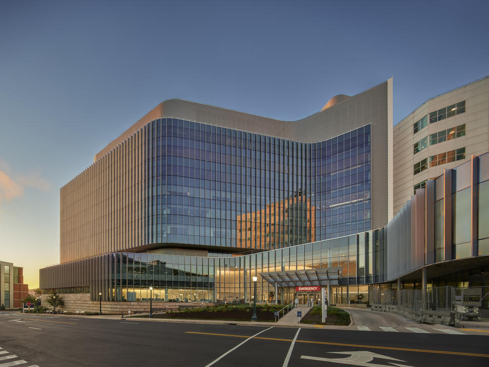 Exterior of a hospital with many reflective windows shot during the sunset. There’s a red entrance sign with white letters that says “Emergency”.