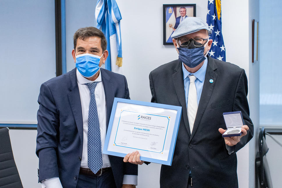 Two men in suits stand next to each other in an office space. Both men wear surgical masks. The man on the right, Enrique Mesri, holds a framed certificate with his name prominently displayed. 