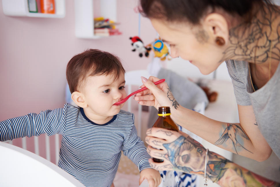 A smiling tattooed woman gives a child a spoonful of dabrafenib. The child is standing in a crib and the background indicates a child’s bedroom.