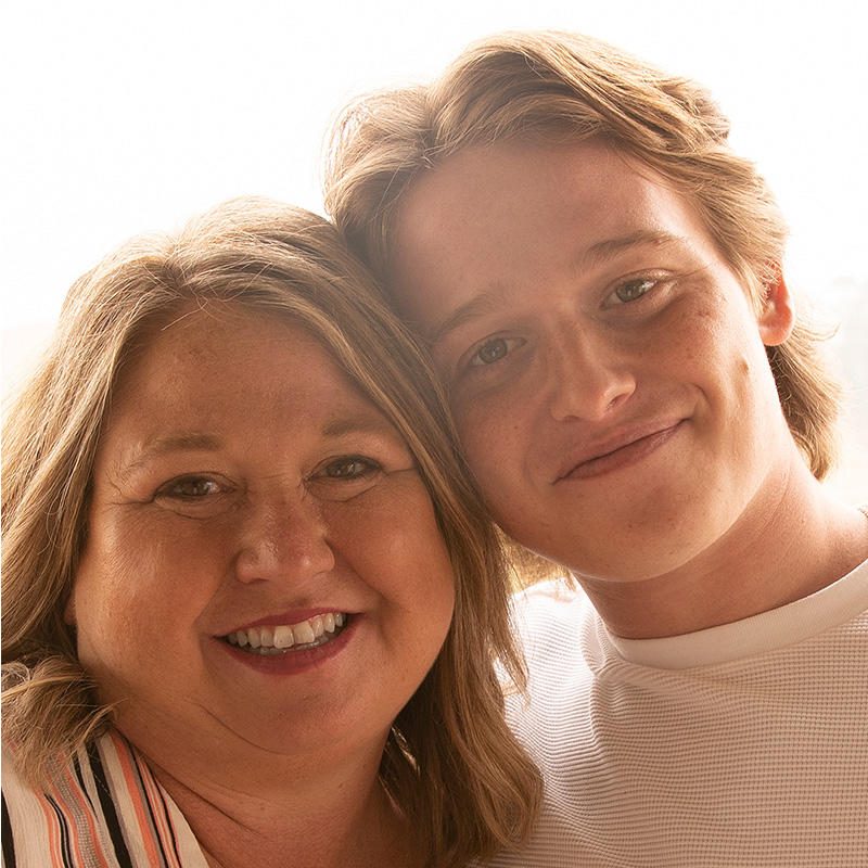 Lisa (a blonde, white woman) stands smiling and leaning into son, Jace (a blonde, white, young adult man).