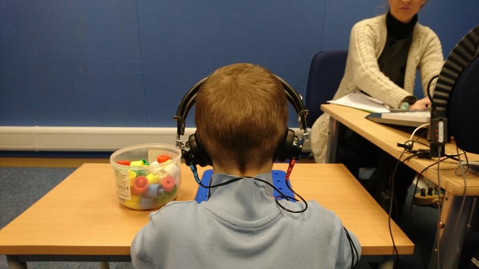 A child wearing headphones sits at a table with colorful plastic pieces in a container in front. In the background, an adult is seated at another table, working with papers and equipment. 