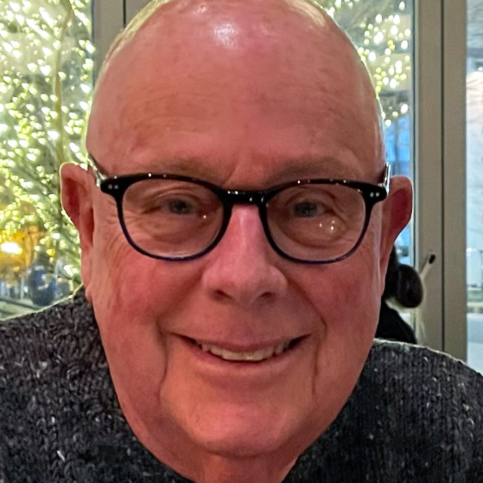 Man with glasses and grey knit sweater smiling at camera while sitting in front of glass doors. Outside are trees covered in white lights.
