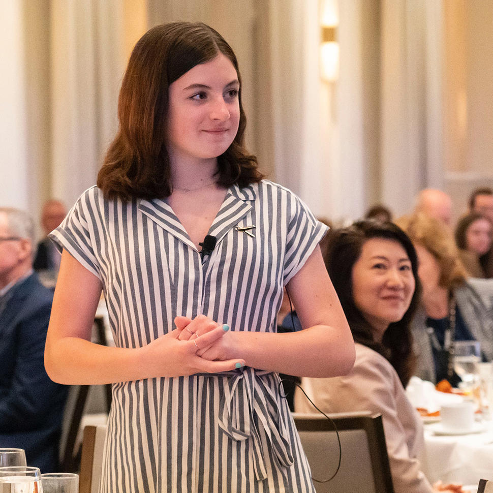 A young woman (Grace) with shoulder-length brown hair is standing and smiling in a room filled with seated people. She is wearing a striped dress with a tie at the waist and has a microphone clipped to her collar. In the background, attendees are seated at tables, engaging in conversation and looking towards the person standing.