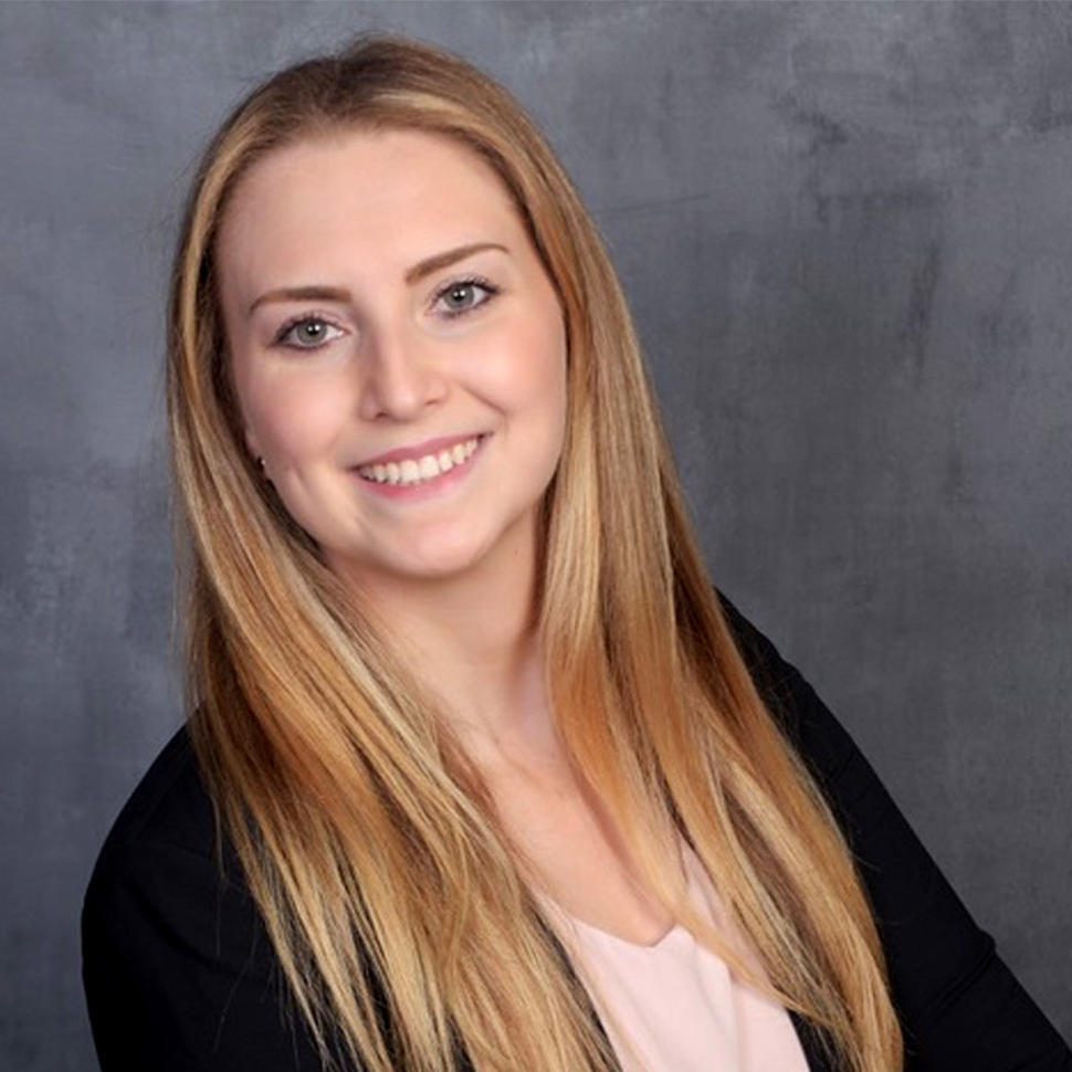Woman with light skin and long, straight blonde hair wearing a pale pink blouse under a dark jacket stands smiling at the camera.