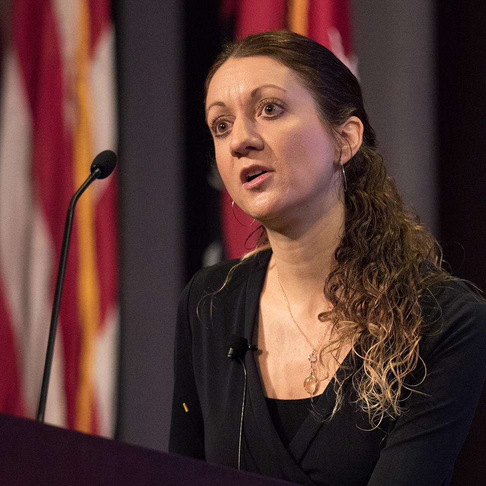Lisa Mirabello is speaking at a podium with a microphone. She has long, wavy hair and is wearing a black outfit. The background features partially visible flags.