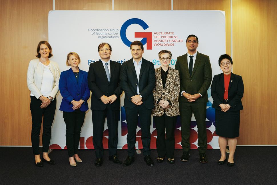 Group of seven individuals stand side by side in front of a backdrop featuring the G7 Cancer logo  and the slogan "Accelerate the Progress Against Cancer Worldwide." They are dressed in formal business attire, including suits and blazers. Behind the backdrop is a wooden wall background.