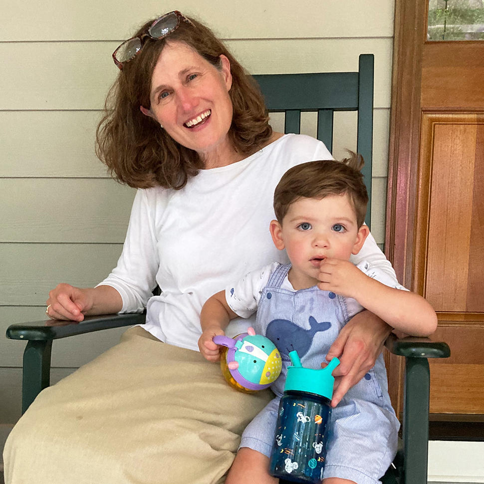 A smiling woman, Dr. Corinne Linardic, with red hair wearing a white shirt and a long, tan skirt sits in a chair with her arm around a child.