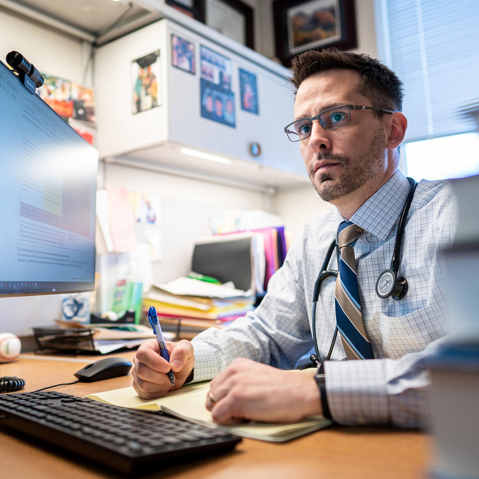 Dr. Matt Ehrhardt, with brown eyes and glasses, sits at a desk, pen in hand, wearing a dress shirt and tie and looking pensively into the distance.