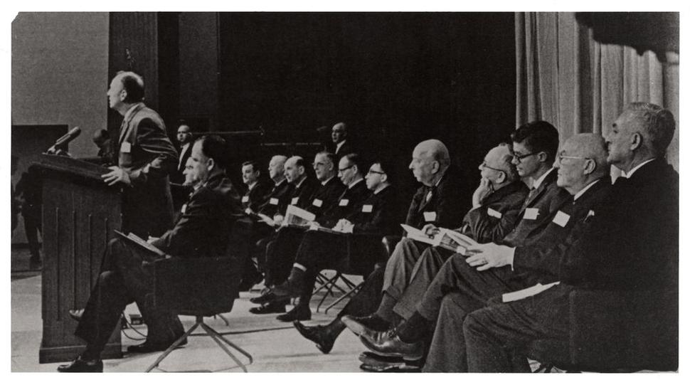 Black and white photo of U.S. Surgeon General Luther Terry addresses press at the release of the 1964 Report on Smoking and Health. Terry stands at a podium as staff and committee sit behind him. 