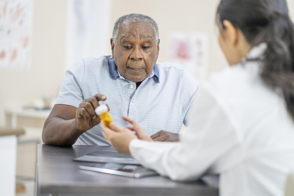 A patient sits across from a doctor holding a prescription pill bottle. The patient and doctor appear to be discussing the medication. 