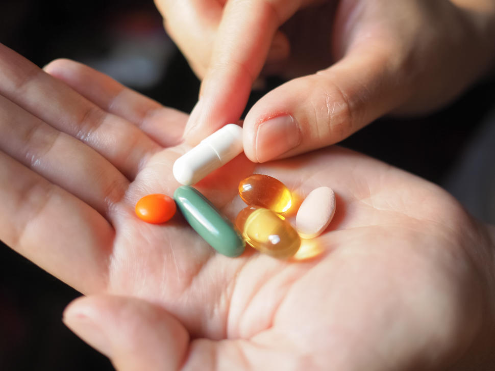 Six pills of various shapes and colors sit in the palm of a person’s hand, with their other hand picking up one of the pills.