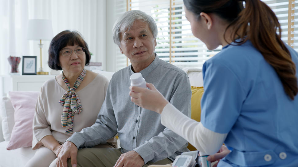 A doctor holds a medicine bottle and speaks with a patient and caregiver seated on a couch.