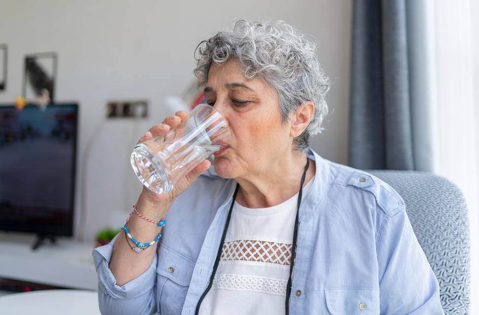 A person with short gray hair drinks water from a tall glass.