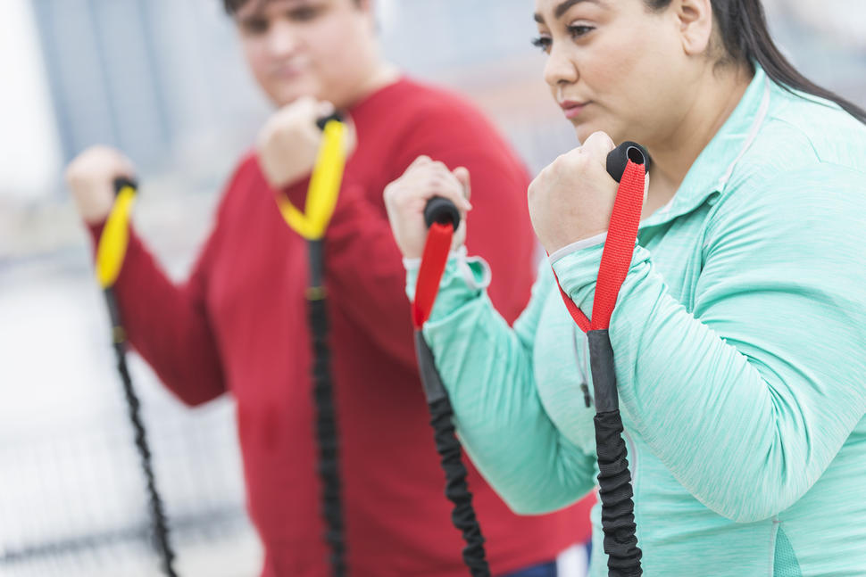 A Hispanic woman in light blue clothes doing curls with exercise bands.