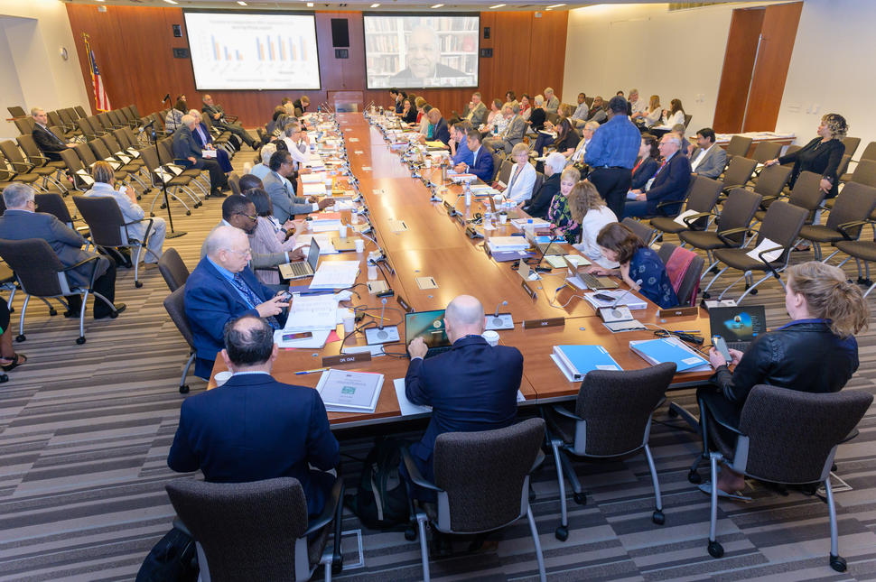 Members of the Board of Scientific Advisors and the National Cancer Advisory Board gather around a table for a joint meeting, with a presentation and remote attendees displayed on screens