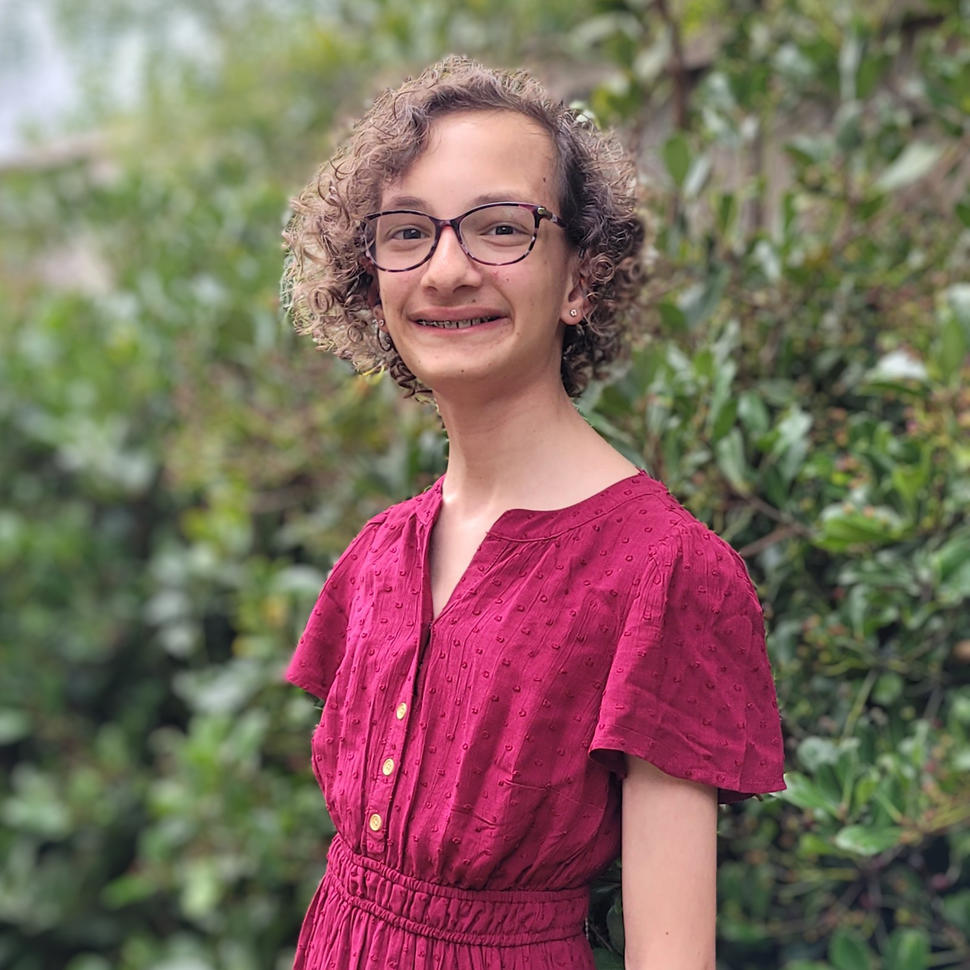 Young girl with curly hair, glasses, and a red dress smiling at the camera.