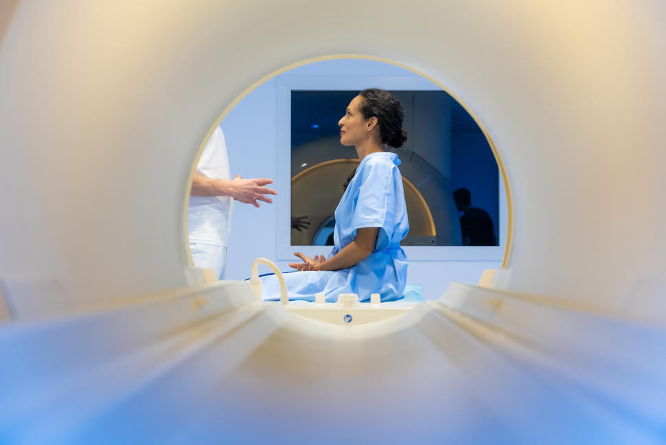 A woman viewed through the opening of an MRI machine. She is seated, wearing a hospital gown, and listening to a health provider who is standing in front of her.