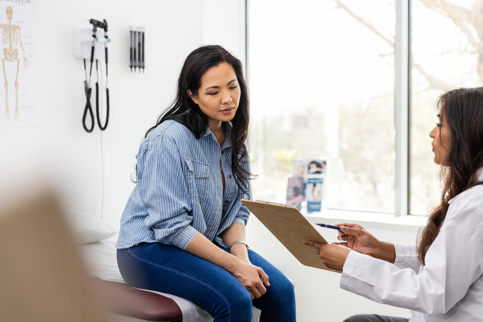 An adult female patient sits on an exam table in a clinic exam room, looking at a medical chart held by a medical provider.