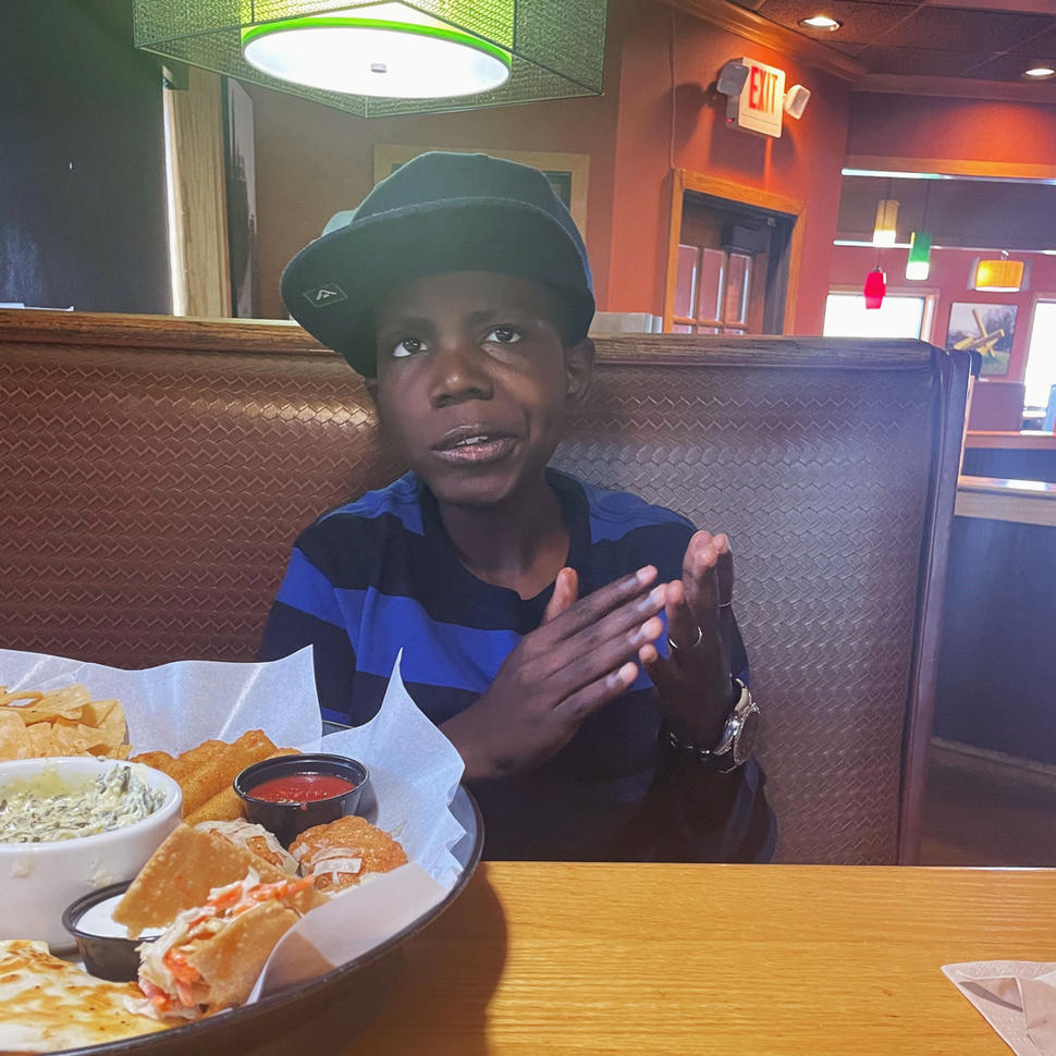 A boy, Cameron, wearing a ball cap and a black and blue striped T-shirt, sits in a restaurant booth and looks toward the camera.
