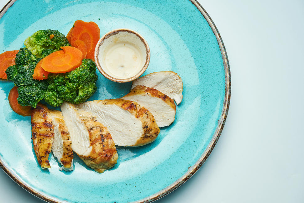 Slices of roasted chicken breast and cooked broccoli and carrots arranged on a blue plate along with a dip in a small bowl.