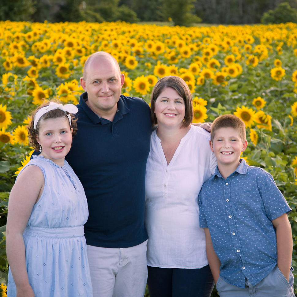 Girl in a blue dress with short hair held back with a headband, standing in a sunflower field with her family.