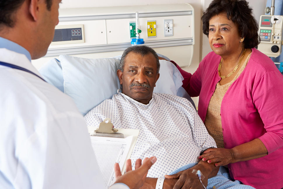 A doctor speaks to a patient in a hospital bed and his caregiver who stands next to the bed with her hand on the patient’s arm.