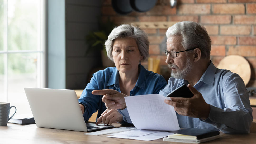Woman and man sit at a table and view a laptop screen as the man holds a printed page in one hand.