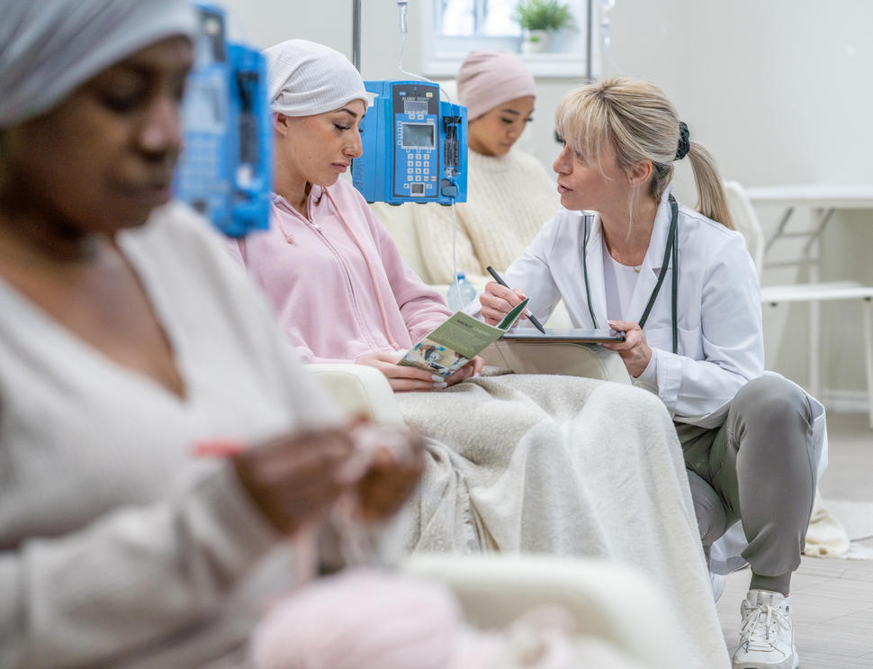 Three women sit in a clinic having IV infusions. A health provider kneels beside and chats with one young woman who is looking at a pamphlet.