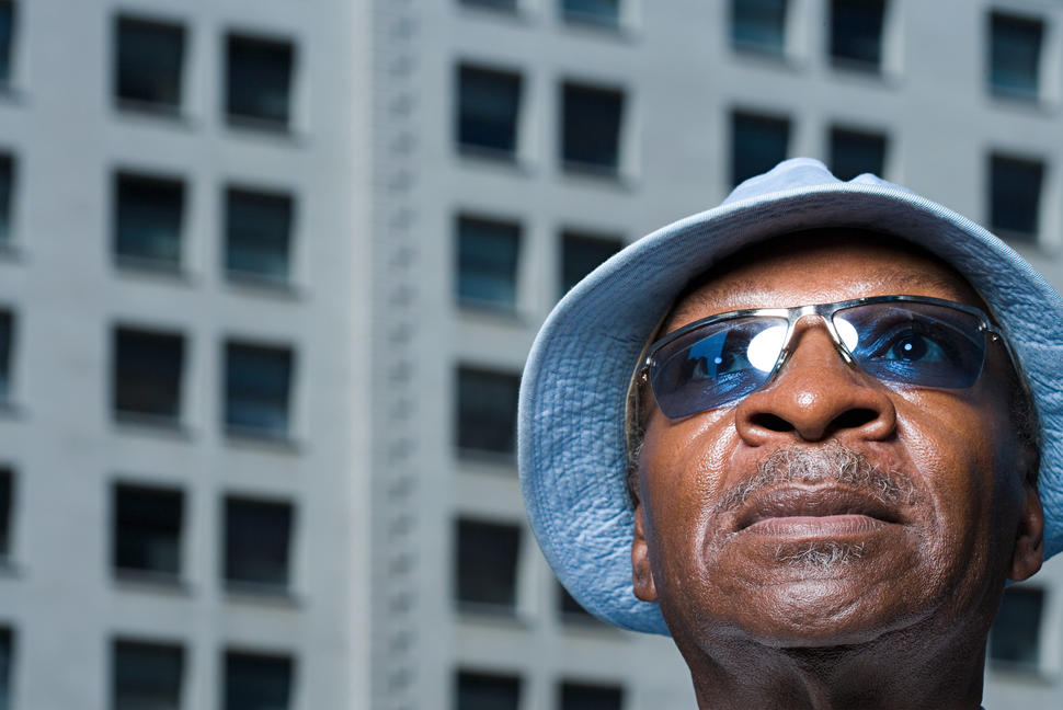 An older dark skinned man wearing a sun hat and sunglasses looks up.