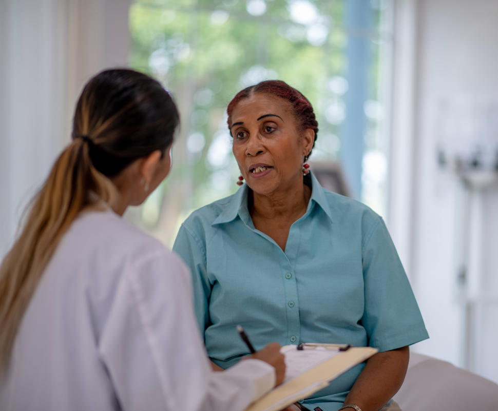 A black woman speaks to a younger woman wearing a white doctor's coat and holding a clipboard and pen.
