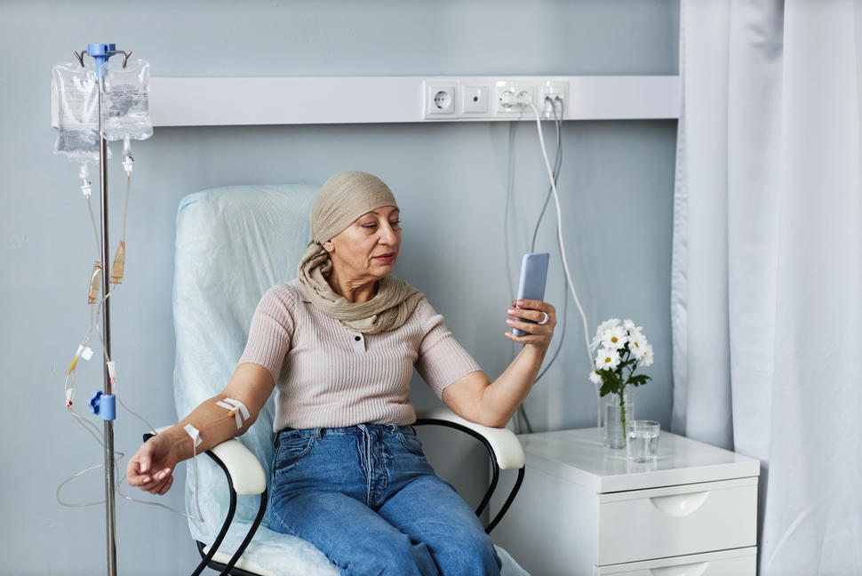An older woman sitting in a chair and looking at a device while receiving an IV treatment.