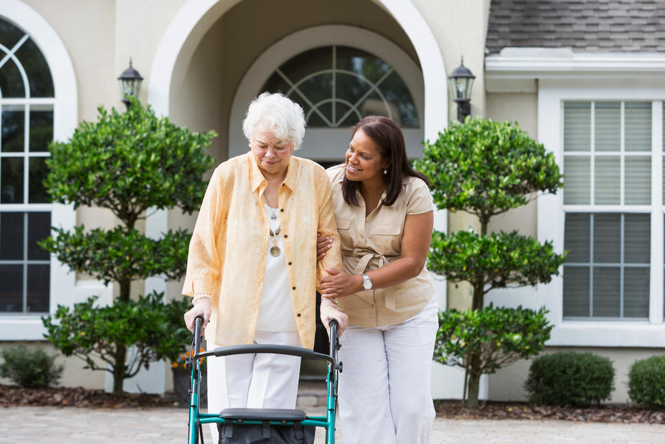Senior woman with walker strolling with middle-aged woman helping her.