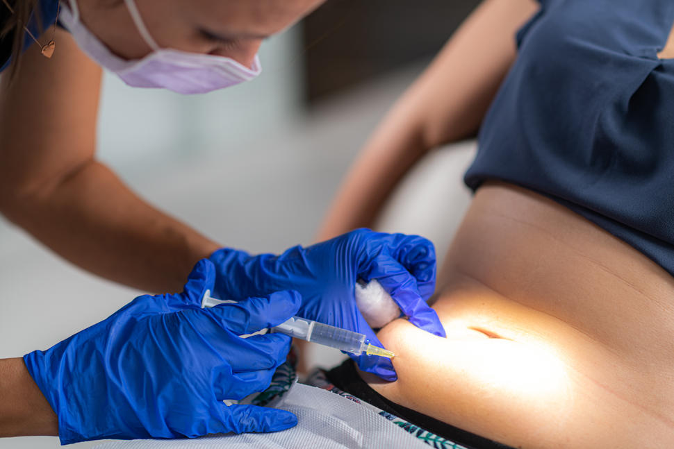 A photograph of a nurse with blue gloves giving a woman an injection of medicine into the lower part of the stomach.
