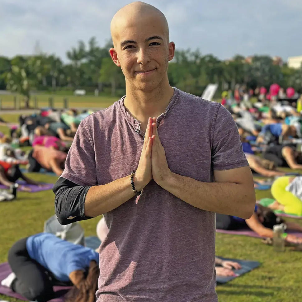 Jace, bald, smiling slightly, hands pressed together in prayer, stands outside in a crowd of people doing yoga.