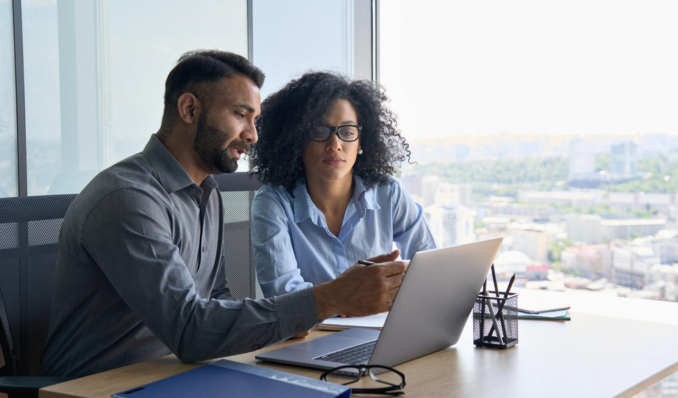 Indian male ceo executive manager mentor giving consultation on financial operations to female African American colleague intern using laptop sitting in modern office near panoramic window.